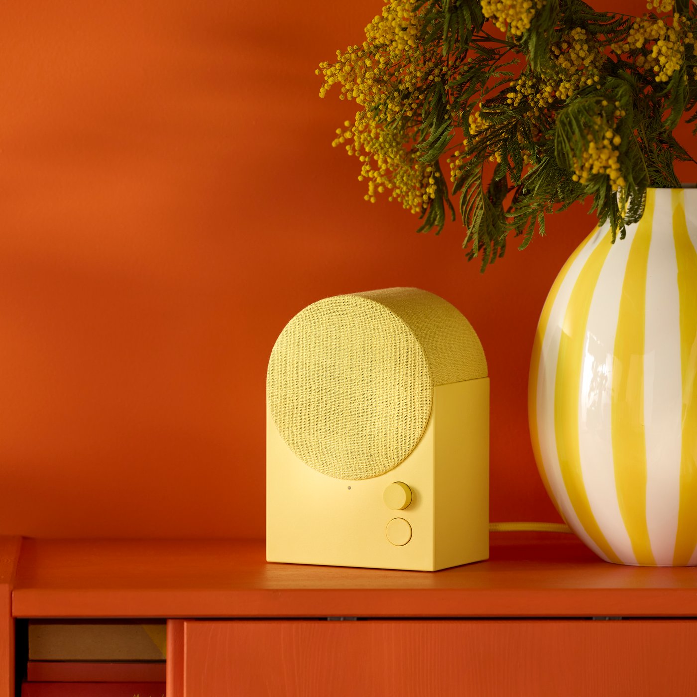 A yellow NATTBAD Bluetooth speaker sits beside a white and yellow vase with flowers on a reddish cabinet with sliding doors.