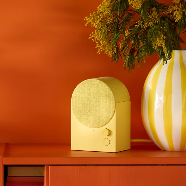 A yellow NATTBAD Bluetooth speaker sits beside a white and yellow vase with flowers on a reddish cabinet with sliding doors.