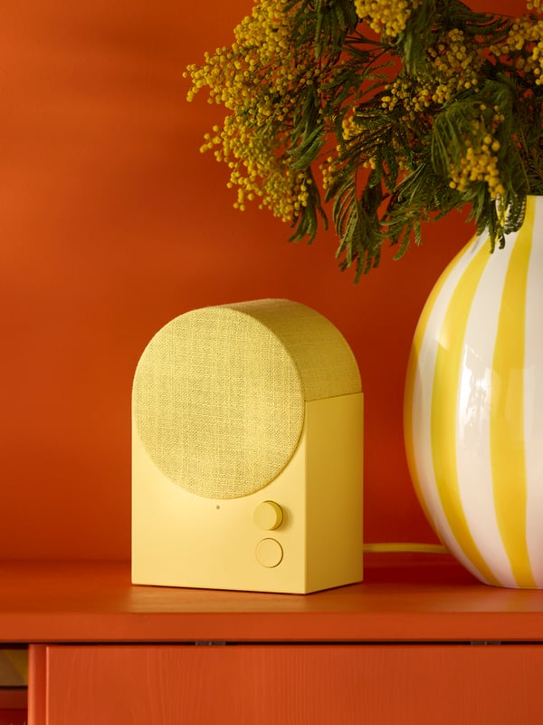 A yellow NATTBAD Bluetooth speaker sits beside a white and yellow vase with flowers on a reddish cabinet with sliding doors.