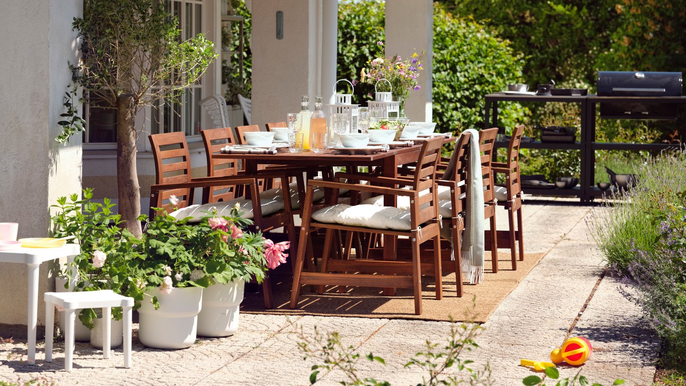 Una mesa de madera ÄPPLARÖ y ocho sillas en una terraza arbolada con una barbacoa GRILLSKÄR cerca y una cadena de iluminación encima.
