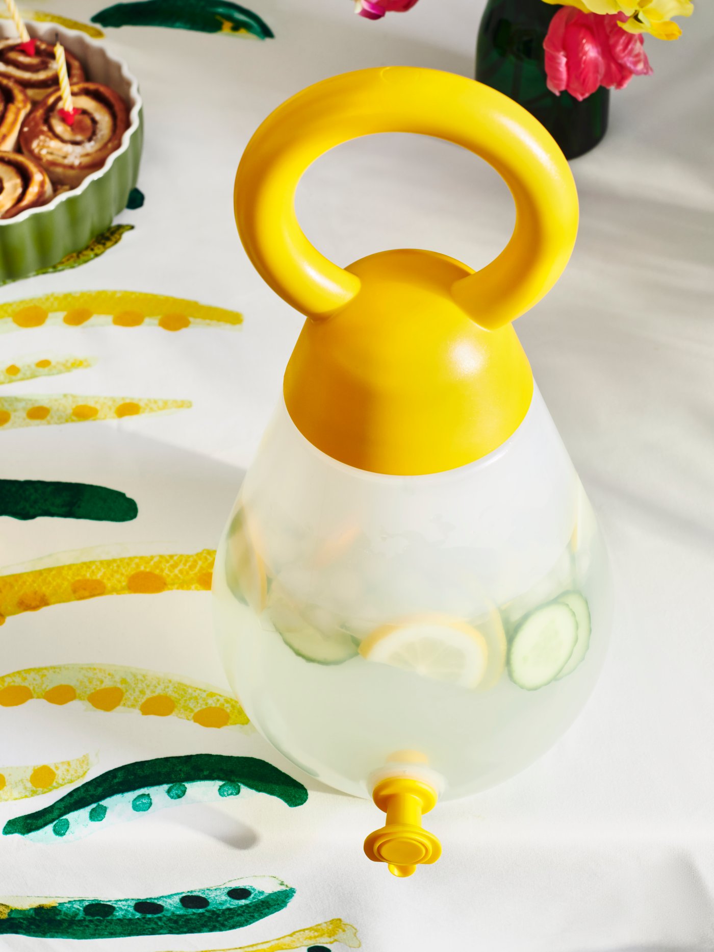 Una caraffa con rubinetto gialla NÄBBFISK con del liquido all'interno su una tovaglia multicolore a fantasia estiva.
