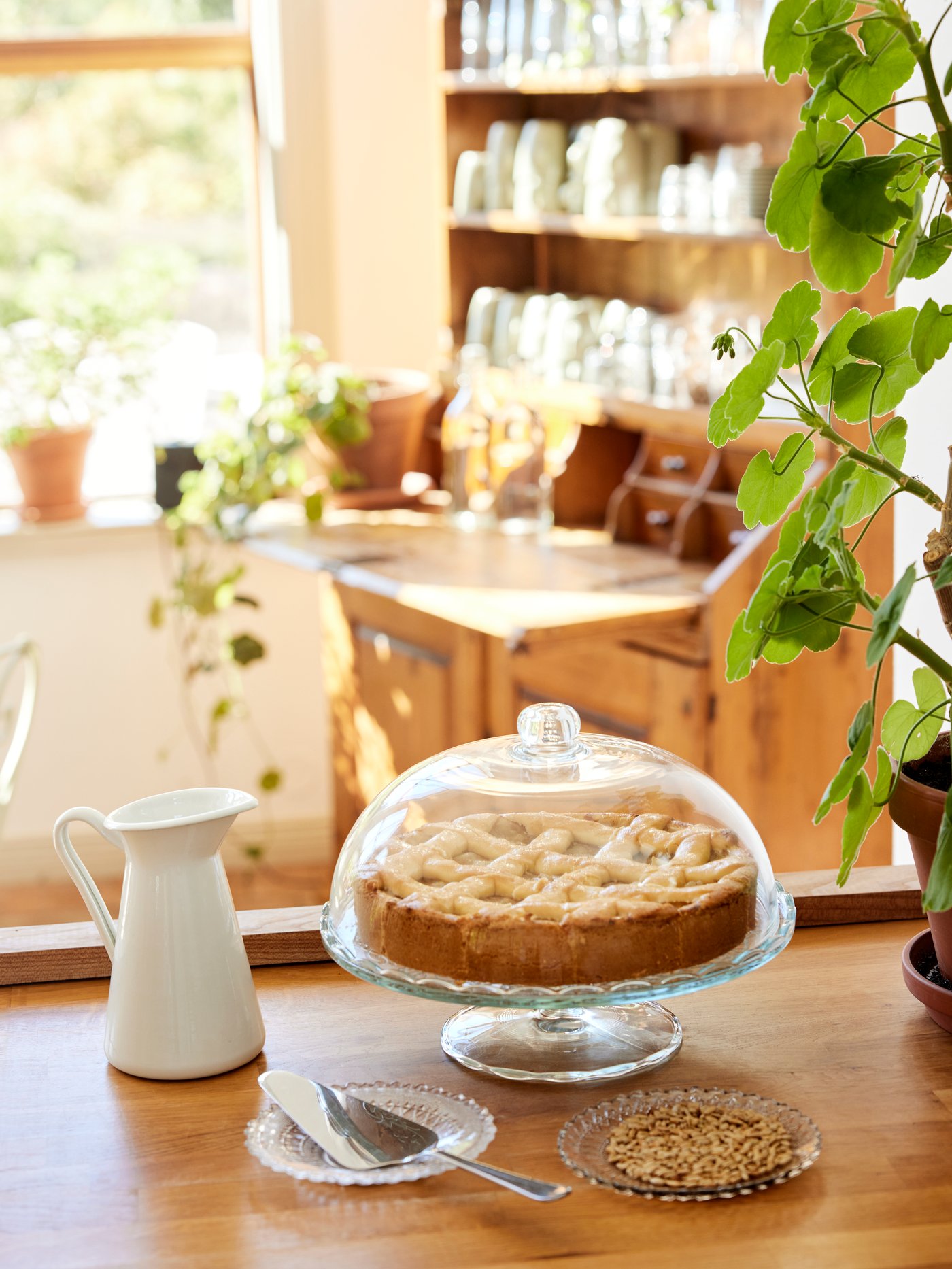 A pie on an ARV BRÖLLOP serving stand with lid beside a white vase, on a wooden counter of a rustic traditional-style café.