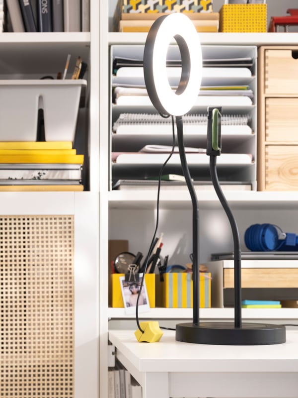 An adjustable LÅNESPELARE ring light with phone holder on a white desk. Office knick-knacks in the background.