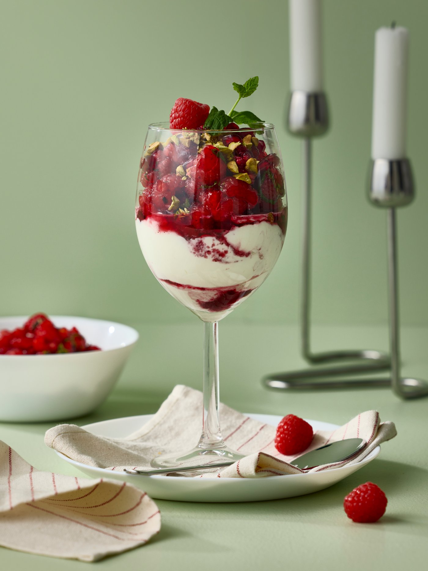 Un bicchiere da vino SVALKA, contenente un dessert con bacche rosse, è appoggiato su un piatto frutta OFTAST bianco con un tovagliolo VIPPSTARR.