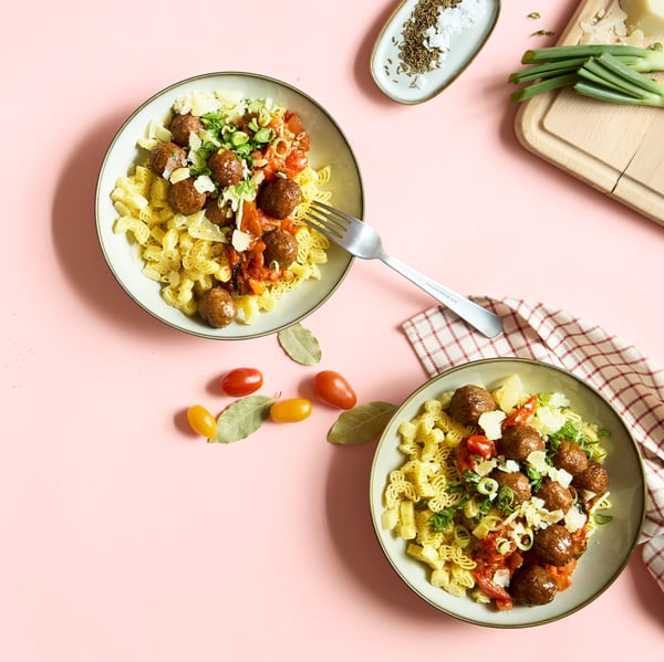 Two deep plates are placed on a pink table and contain a dish of pasta, plant balls, and a sauce of tomato and spinach.