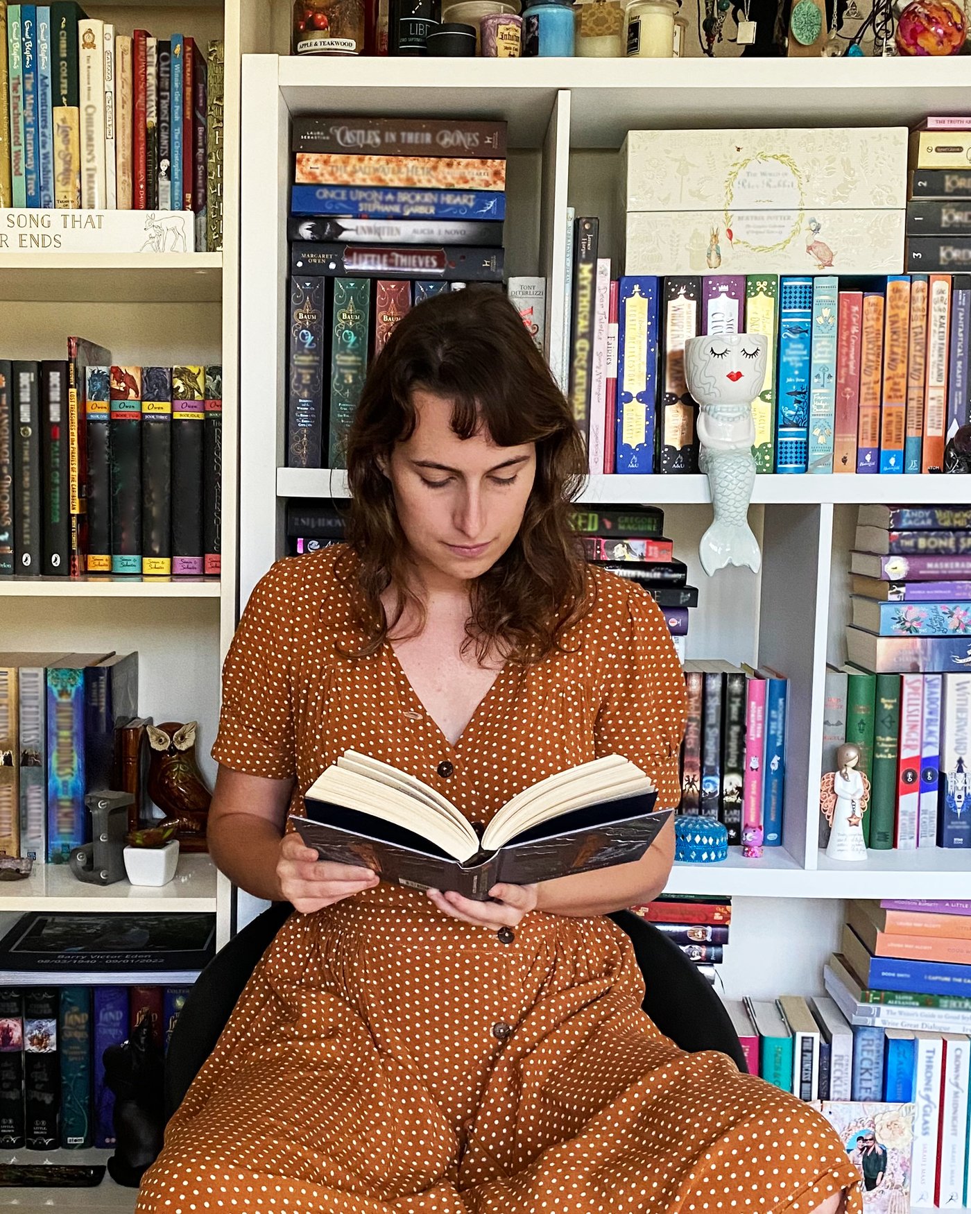 Een vrouw in een lange jurk zit in een stoel en leest een boek. Aan de wand achter haar hangt een schap dat vol staat met boeken.