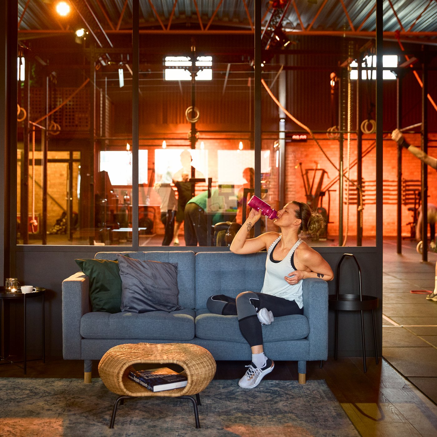 A woman in a crossfit gym drinking water on a grey sofa