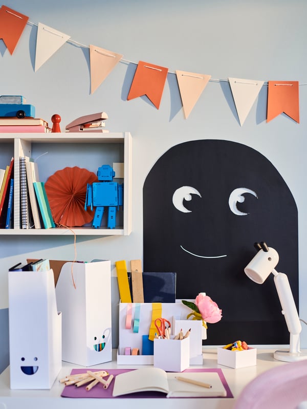 A child's desk space with white TJABBA magazine files, a white KRUX work lamp and a wide range of school supplies.