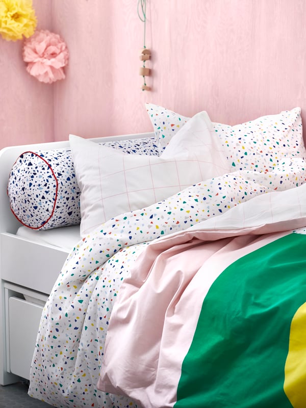 Una cama infantil SLÄKT con almacenamiento inferior, cubierta por sábanas y cobertores MÖJLIGHET en tonos rosado claro y verde. 