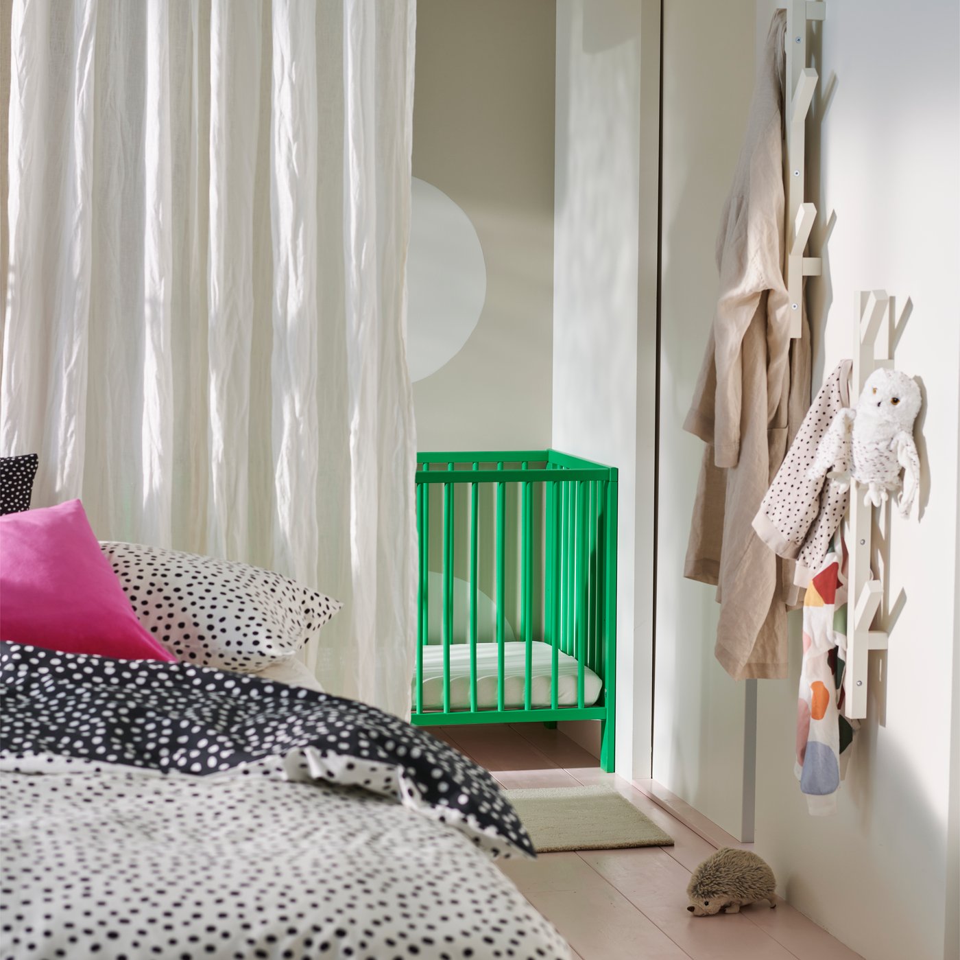 En sida av en VIHALS säng bäddad med BJÖRKGRÅMAL sängkläder framför en vit DYTÅG gardin och en grön GULLIVER spjälsäng.