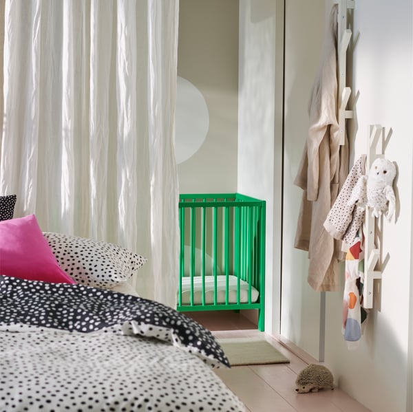 One side of a VIHALS bed covered in BJÖRKGRÅMAL bed linen in front of a white DYTÅG curtain and a green GULLIVER cot.