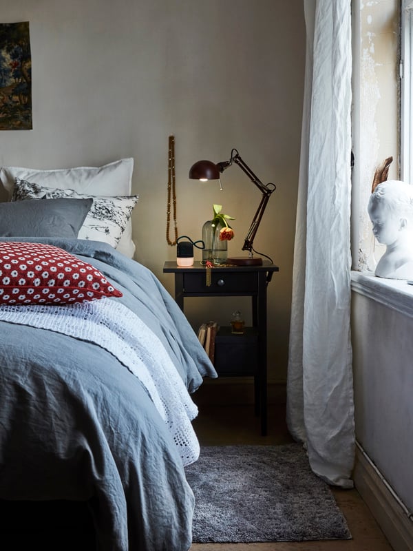 A section of a bed with gray ÄNGSLILJA bed linen and a patterned cushion beside a black-brown HEMNES bedside table.