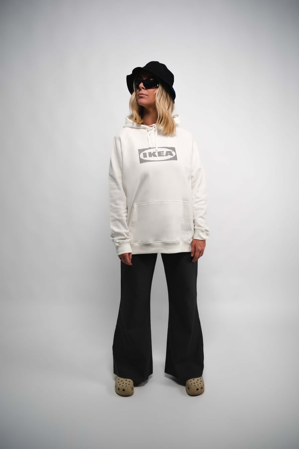 Modell i svart hatt, vit hoodie med IKEA-logotyp, svarta utsvängda byxor och beiga skor – en minimalistisk streetwear-look från IKEAs AURTIENDE kollektion.