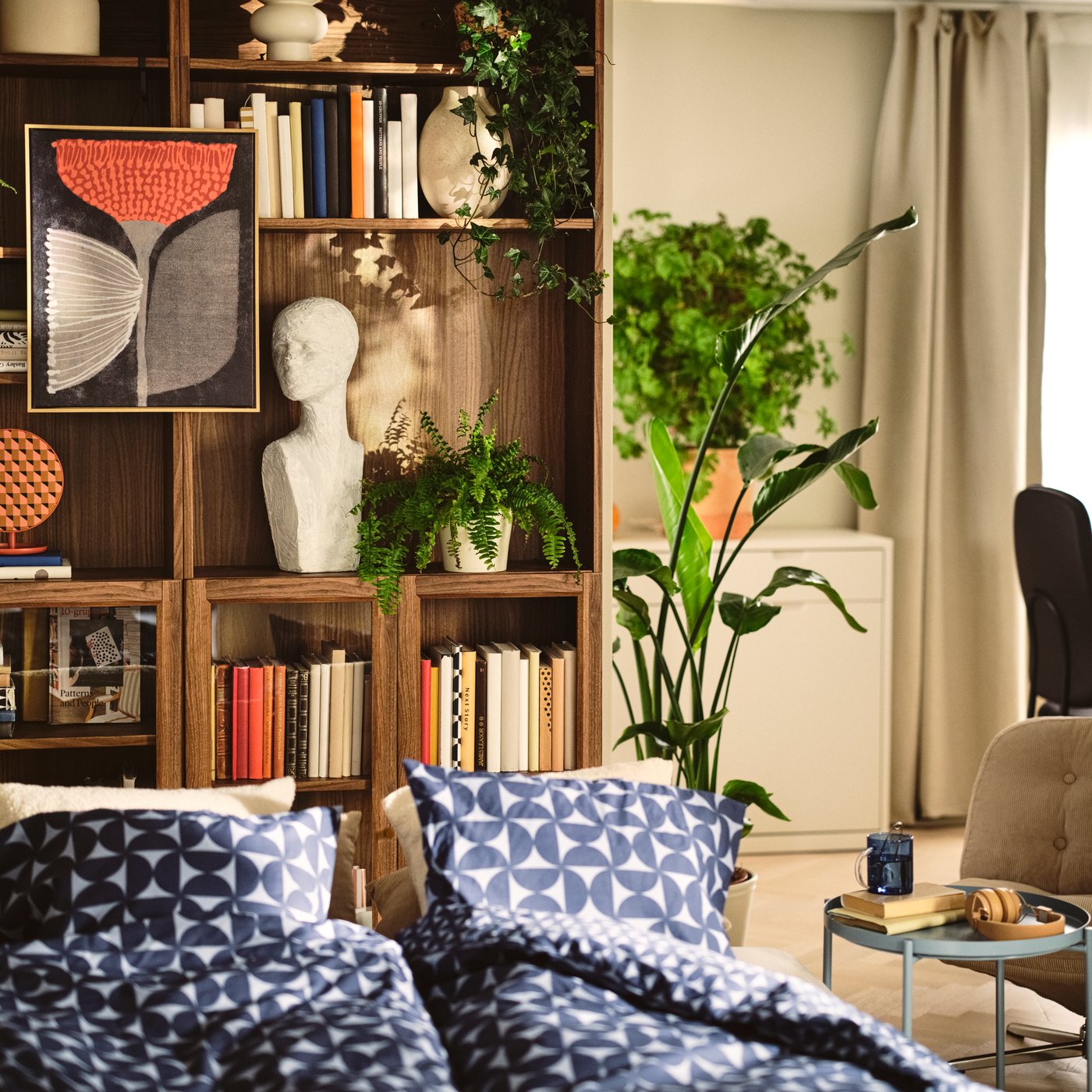 A living room with a beige SÖDERHAMN 3-seat sofa-bed, upholstered in Kelinge beige, surrounded by cushions and plants.