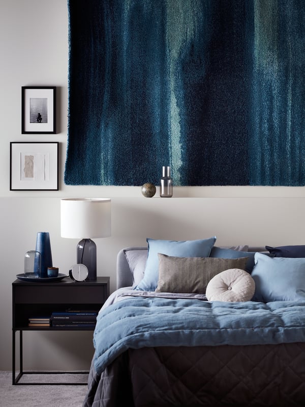 A TONVIS table lamp on a black VIKHAMMER bedside table by a SLATTUM bed with KOPPARBLAD bed linen and a blue rug on the wall.