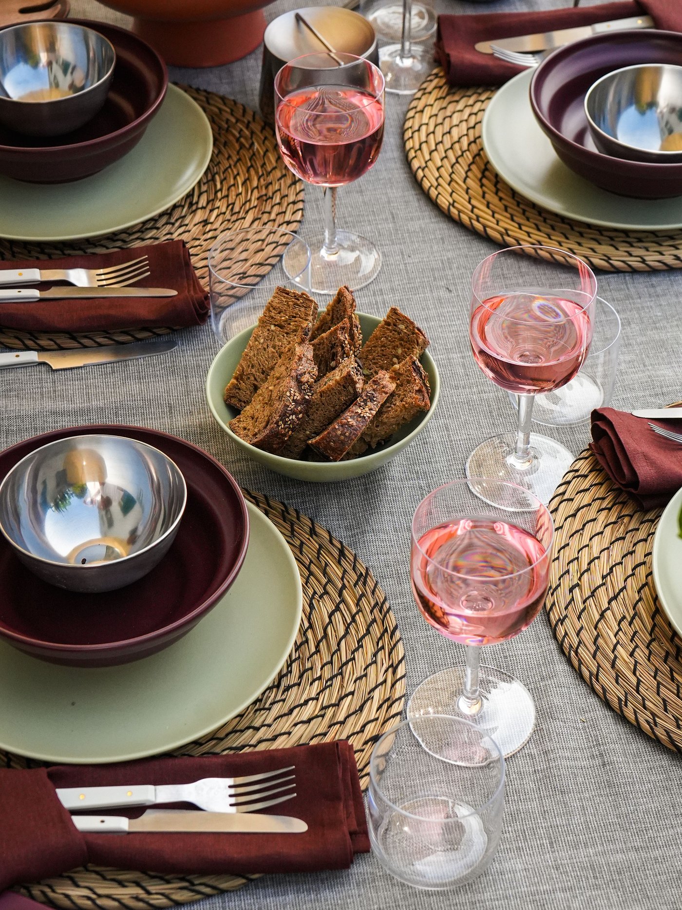 Une table dressée avec des sets de table tressés « LÄTTAD », des assiettes vert clair « FÄRGKLAR », des bols rouge-brun « SPJUTLILJA », des couverts et du vin rosé. Du pain, du fromage, des olives.