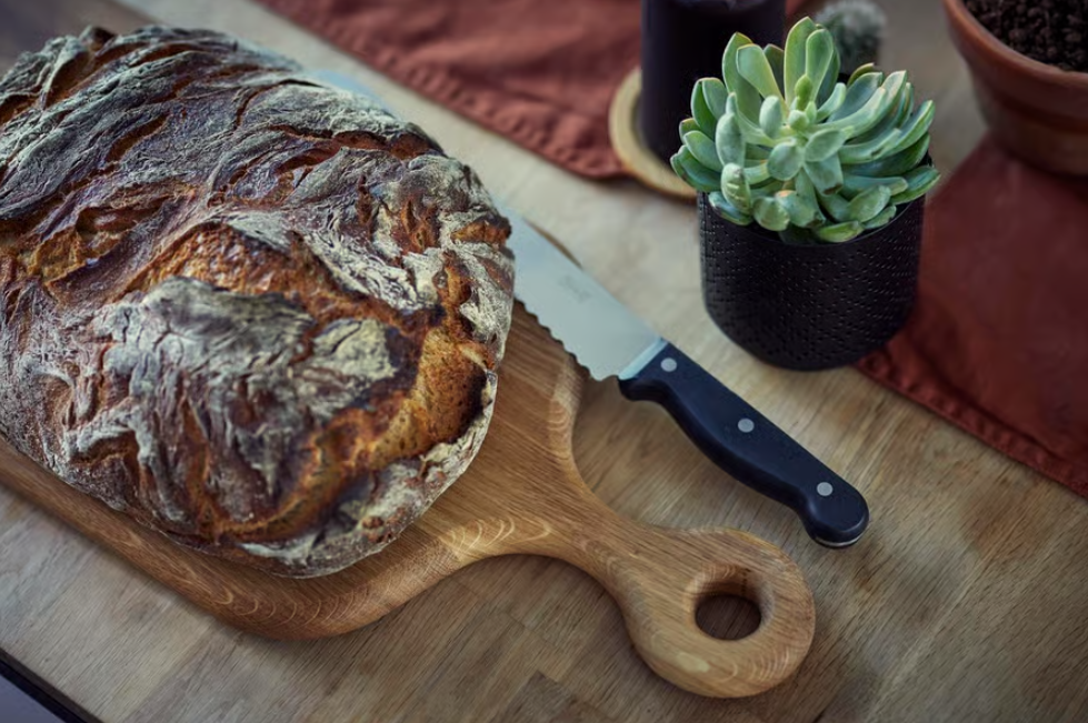 Laib Brot auf Holzbrett mit Messer, daneben eine kleine Sukkulente und ein rotes Platzset auf Holztisch.