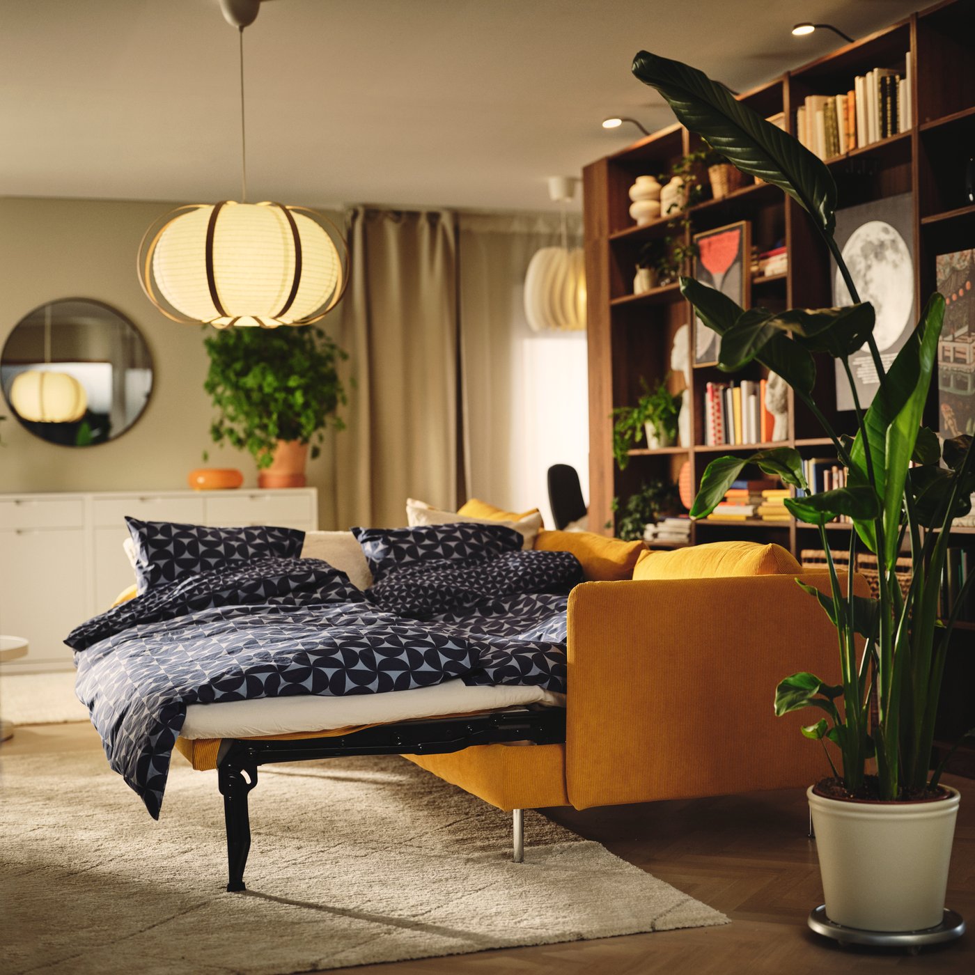 A living room with a dark yellow SÖDERHAMN 3-seat sofa-bed surrounded by plants and light curtains.