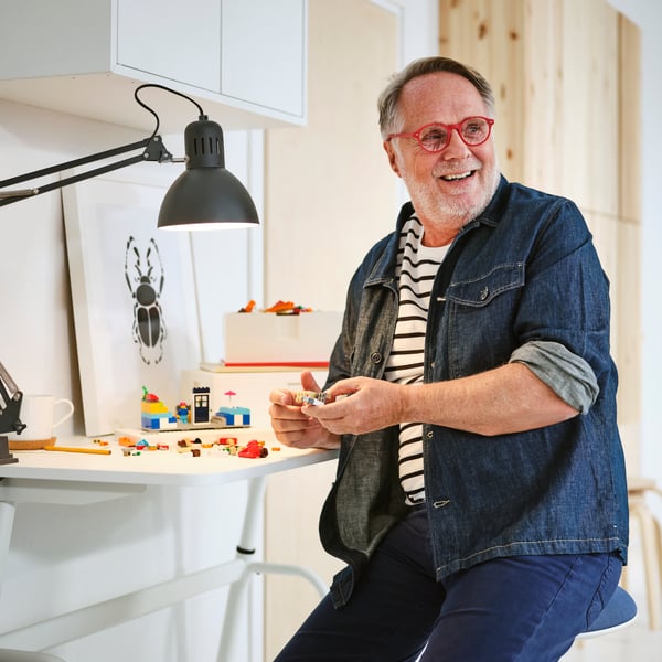 En person i jeansskjorta och jeans på ett NILSERIK sitt-/ståstöd bygger med BYGGLEK LEGO klossar vid ett vitt GLADHÖJDEN skrivbord.