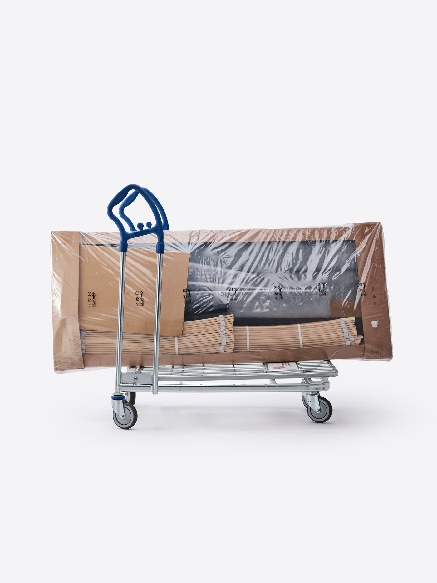 Rám postele SLATTUM, zabalený naplocho v plastu a kartonu, leží na nákupním vozíku IKEA.