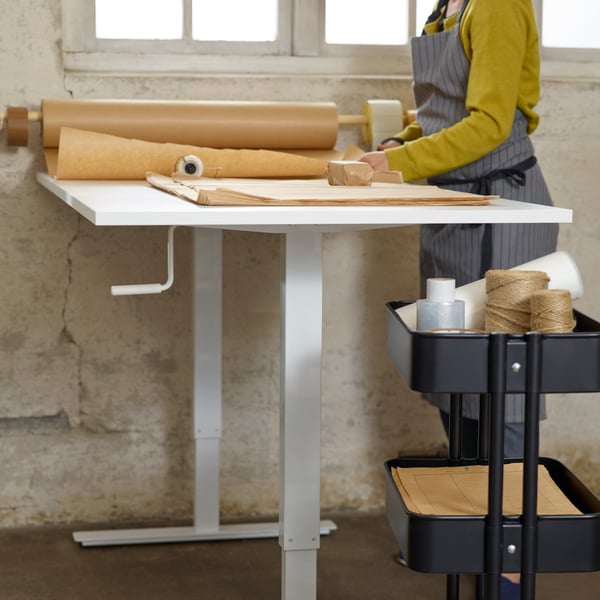 Uma pessoa de pé junto a uma mesa de trabalho em branco, a ajustar alguns rolos de materiais de trabalho castanhos, e um carrinho preto por perto.