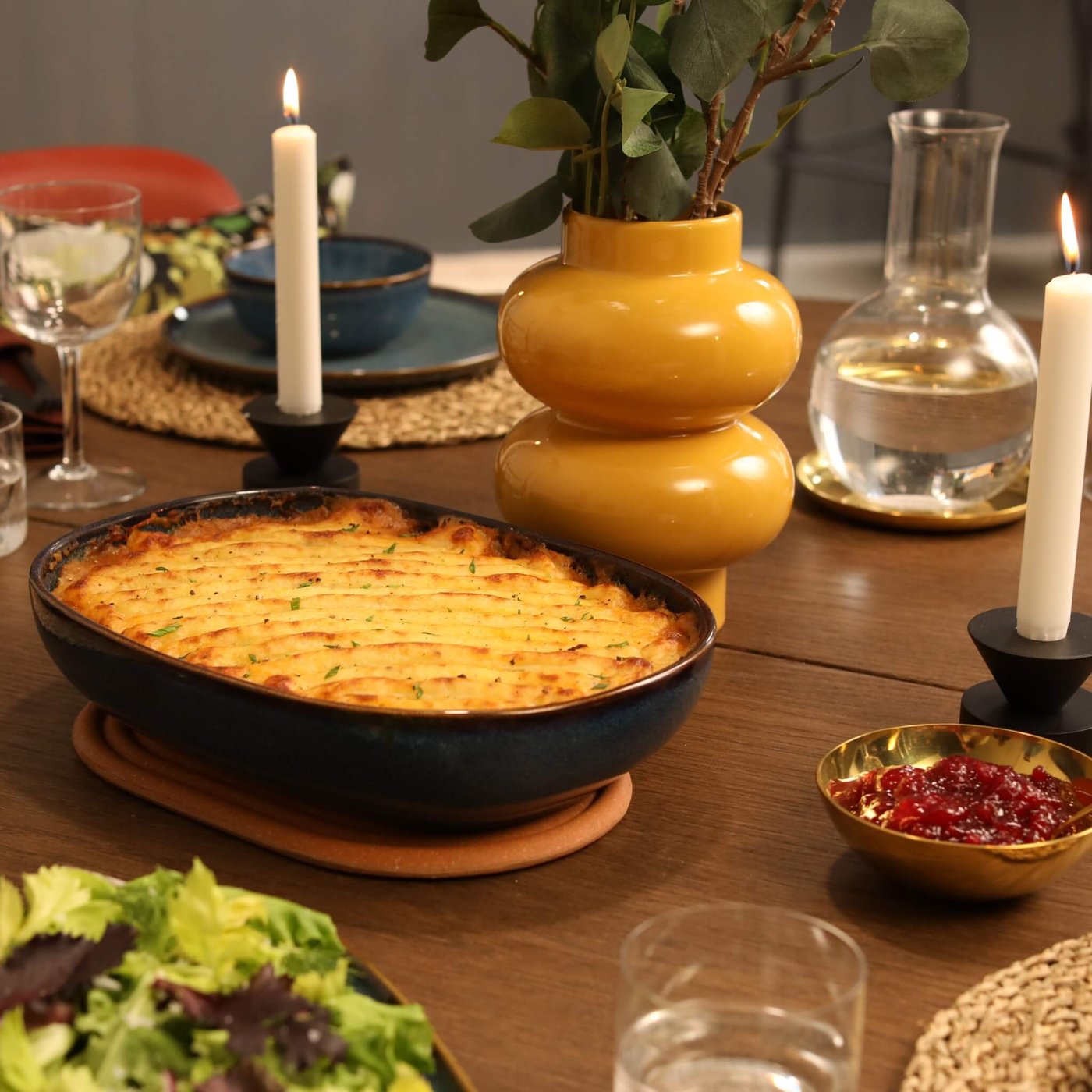 Shepherd's pie on a set dinner table with candles, salad, lingonberry sauce and a yellow vase.