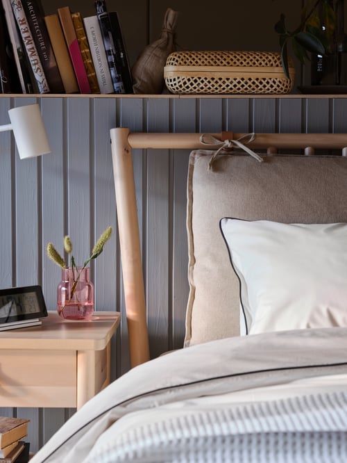 Upper left side of a beige bed frame with a duvet set. Next to the bed is a bedside table with an alarm clock and pink vase.