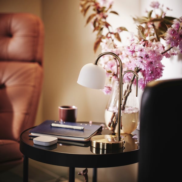 A side table with a notebook and pen, pink flowers in a vase and TVÅMASTAD LED portable lamp.