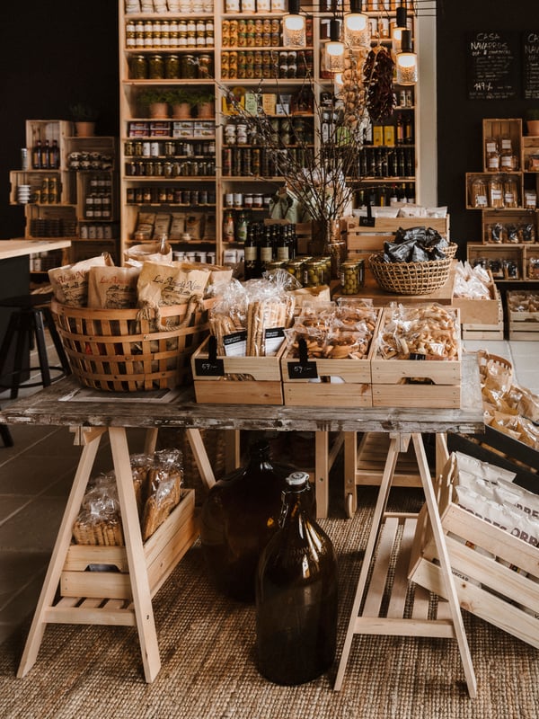 Rustik butik med trähyllor och bord fyllda med varor som flaskor, burkar och korgar. Mysig atmosfär med varm belysning och välorganiserade produkter.