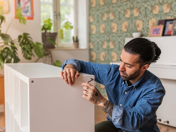A person is assembling a white piece of furniture in a warmly decorated room with plants and artworks.