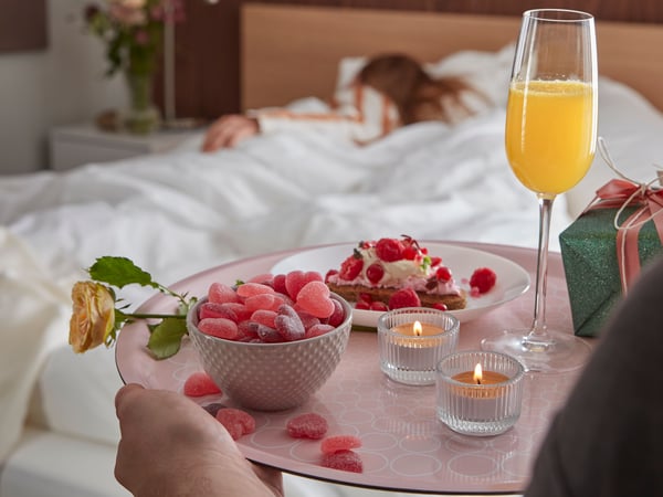 En person kommer med frukost på sängen på en rosa KEJSERLIG bricka till en annan person som ligger och sover i en säng.