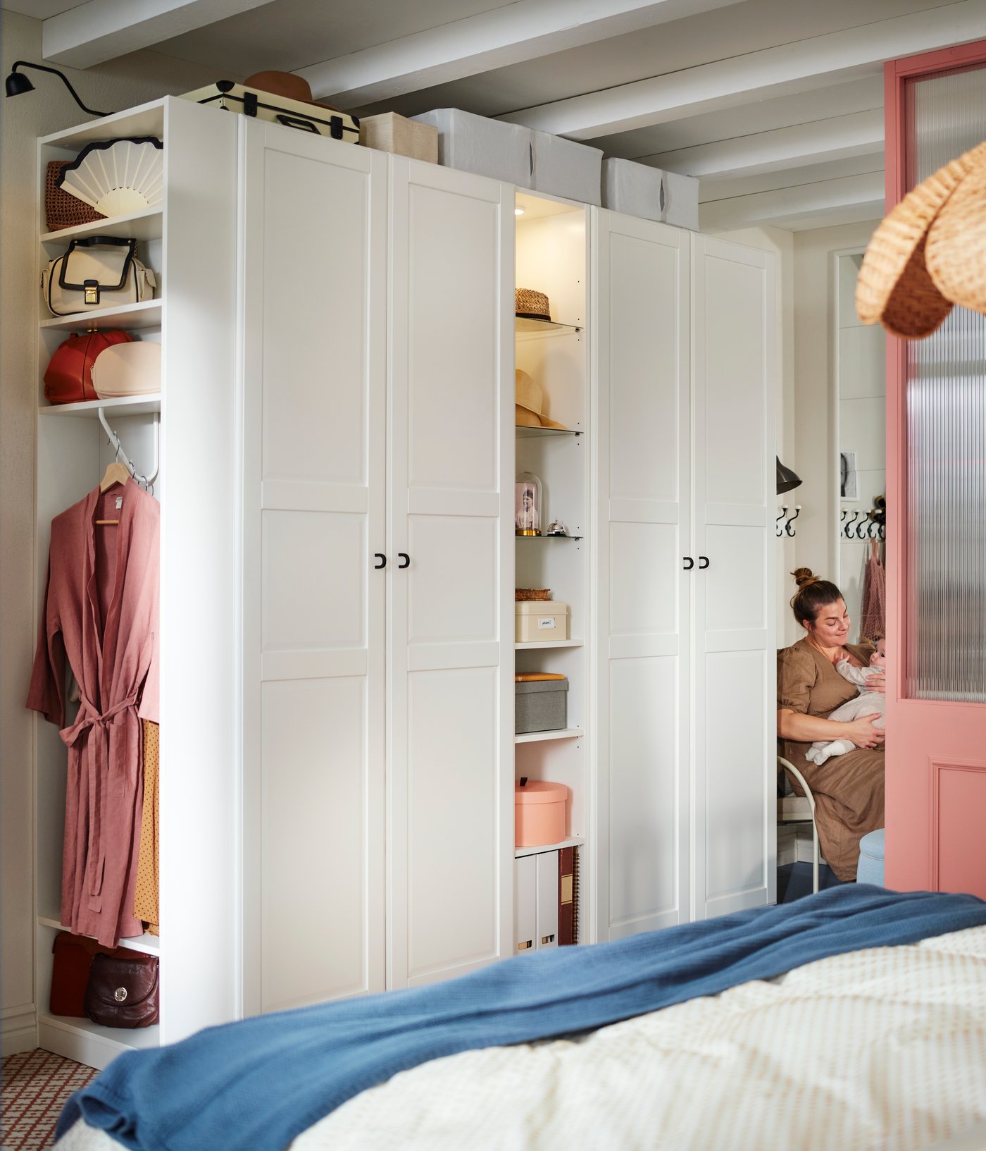 In una camera da letto tradizionale, una donna con un bambino in braccio è seduta accanto a un guardaroba PAX/TYSSEDAL bianco con un elemento supplementare terminale e un elemento supplementare centrale.