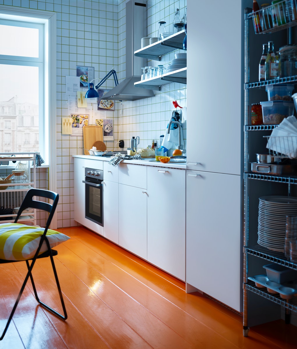 白いキャビネットとオレンジ色の床のキッチン。壁に調理器具が掛かり、窓際には椅子とクッションが置かれている。