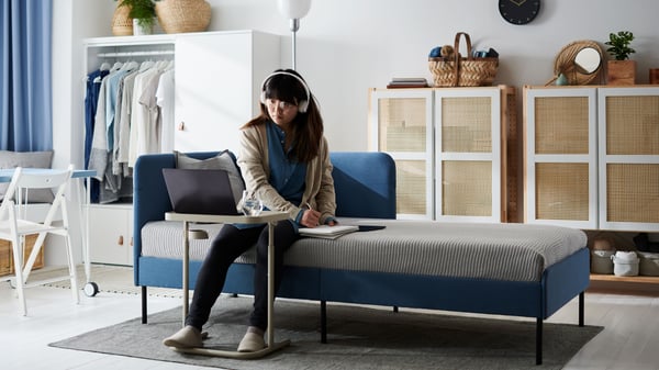 A young woman with headphones sits on the side of a BLÅKULLEN bed, focusing on a laptop placed on a BJÖRKÅSEN laptop stand.