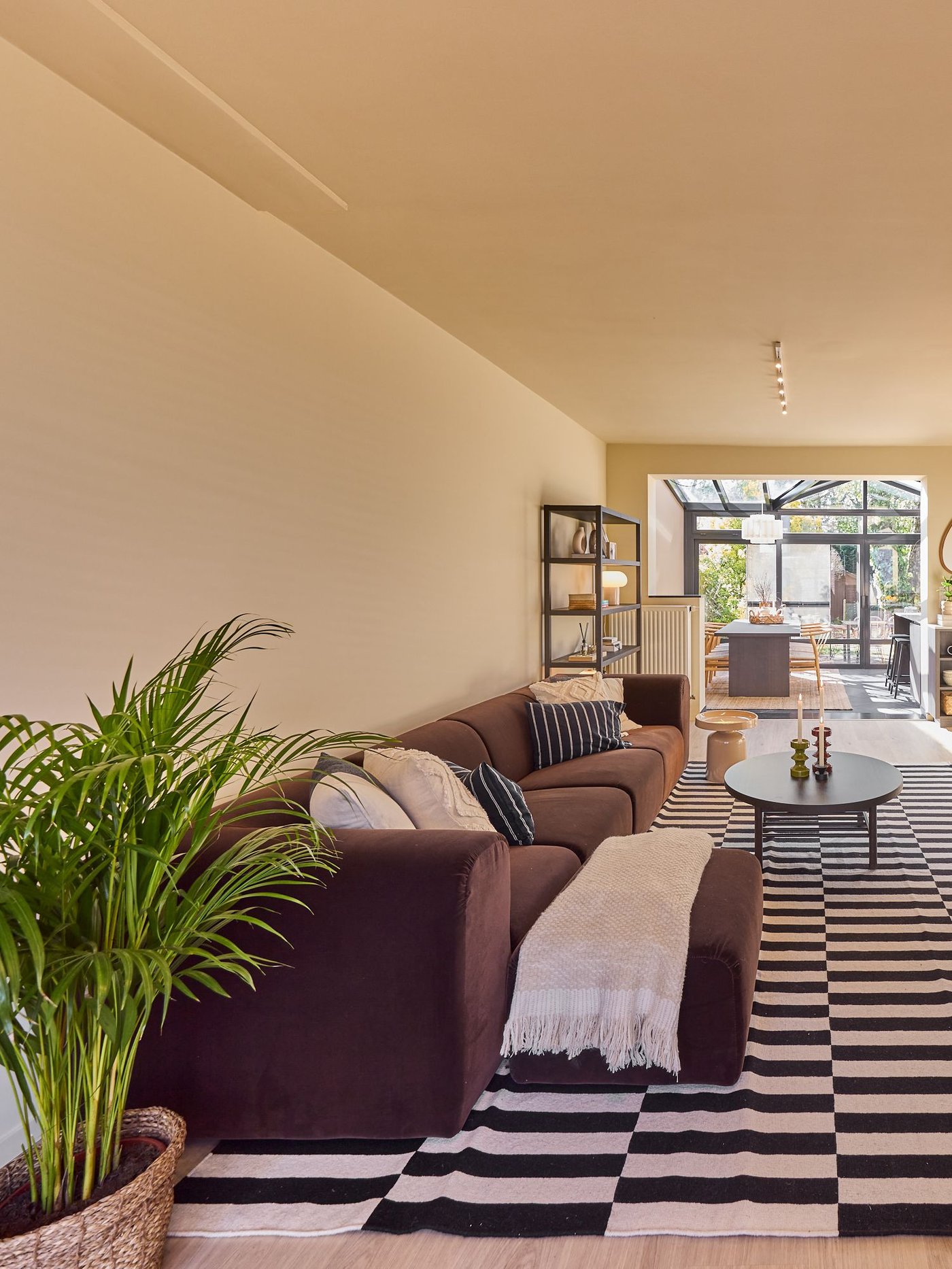 Woonkamer met bruine zetel, gestreept tapijt en salontafel, uitkijkend op een lichte eetruimte en tuin erachter.