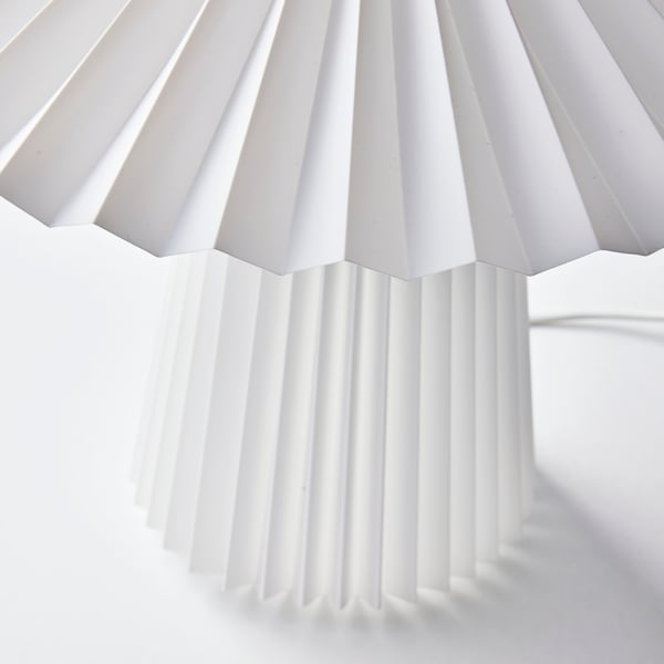The top of a white KUDDLAVA table lamp, with light playing in the pleats of the circular shade around a central birch knob.