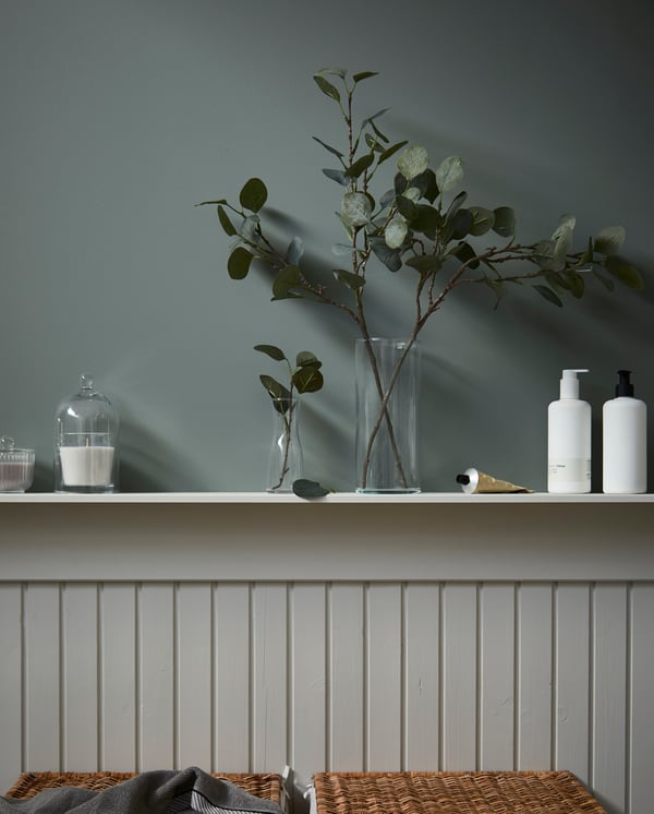 A half-panel wall with a shelf where a glass dome covers a candle and artificial olive twigs are displayed in glass vases.
