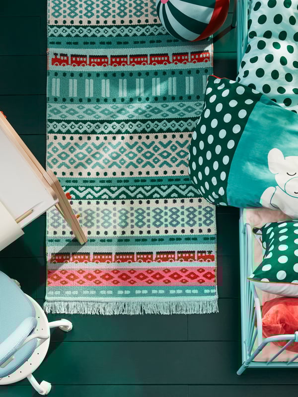 A KÄPPHÄST rug and a KÄPPHÄST soft toy shaped like a ball lie on a floor beside a bed with colourful bed linen.