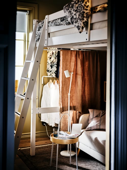 Schlafzimmer mit einem STORÅ Hochbett, einem Sofa und einem BURVIK Beistelltisch darunter sowie einer Kleiderstange seitlich, die durch Stoff verdeckt ist.