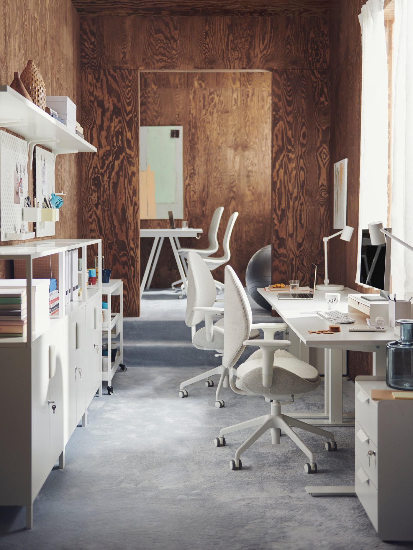 A small office with dark brown wooden walls, white TROTTEN desks, TROTTEN cabinets, and LÅNGFJÄLL chairs.