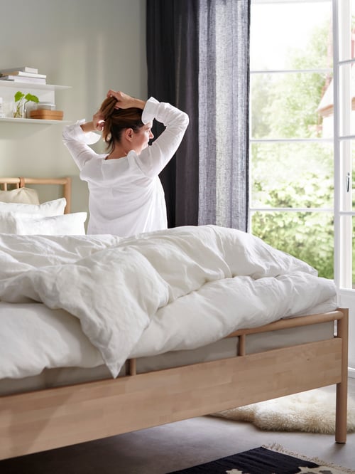 A woman sitting on a BJÖRKSNÄS bed facing a window with dark grey curtains. The bed has white ÄNGSLILJA bedlinen on it.