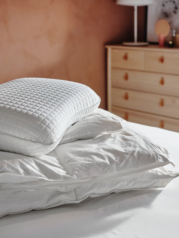 A FJÄLLARNIKA duvet and a KLUBBSPORRE ergonomic pillow are piled-up together on a bed in a sun-filled room.