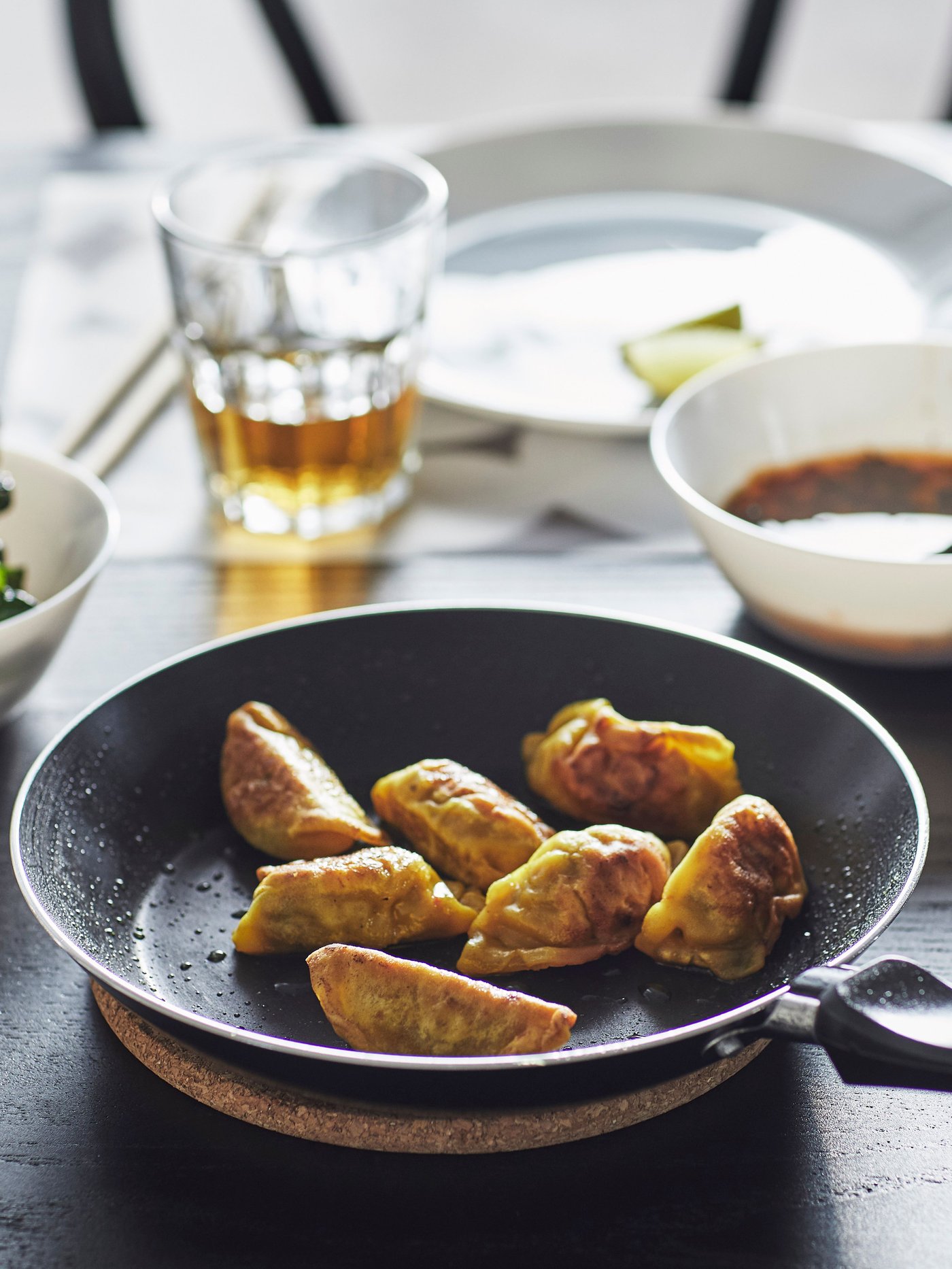TAGGHAJ frying pan with pan-fried gyozas on a HEAT pot stand on the dining table, next to various dinnerware and glasses. 
