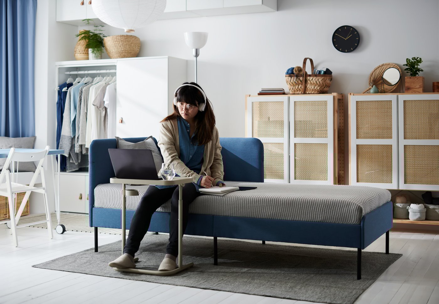 A young woman with headphones sits on the side of a BLÅKULLEN bed, focusing on a laptop placed on a BJÖRKÅSEN laptop stand.