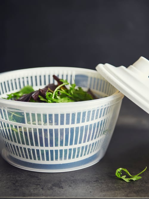 Gros plan d’un comptoir de cuisine sombre avec des murs sombres et une essoreuse à salade avec un bol en plastique transparent, une passoire et un couvercle blancs.