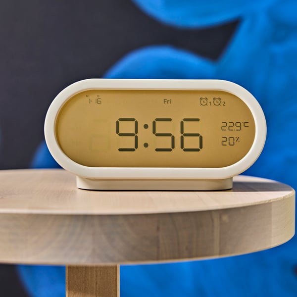 Primer plano de un reloj despertador RÖDSTRÄFSE color beige sobre una mesa auxiliar de madera, mostrando la hora, la temperatura y la humedad.