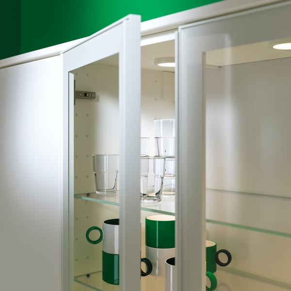 A wall cabinet has one of the glass doors open and there are glasses and green and white mugs stored on glass shelves inside.