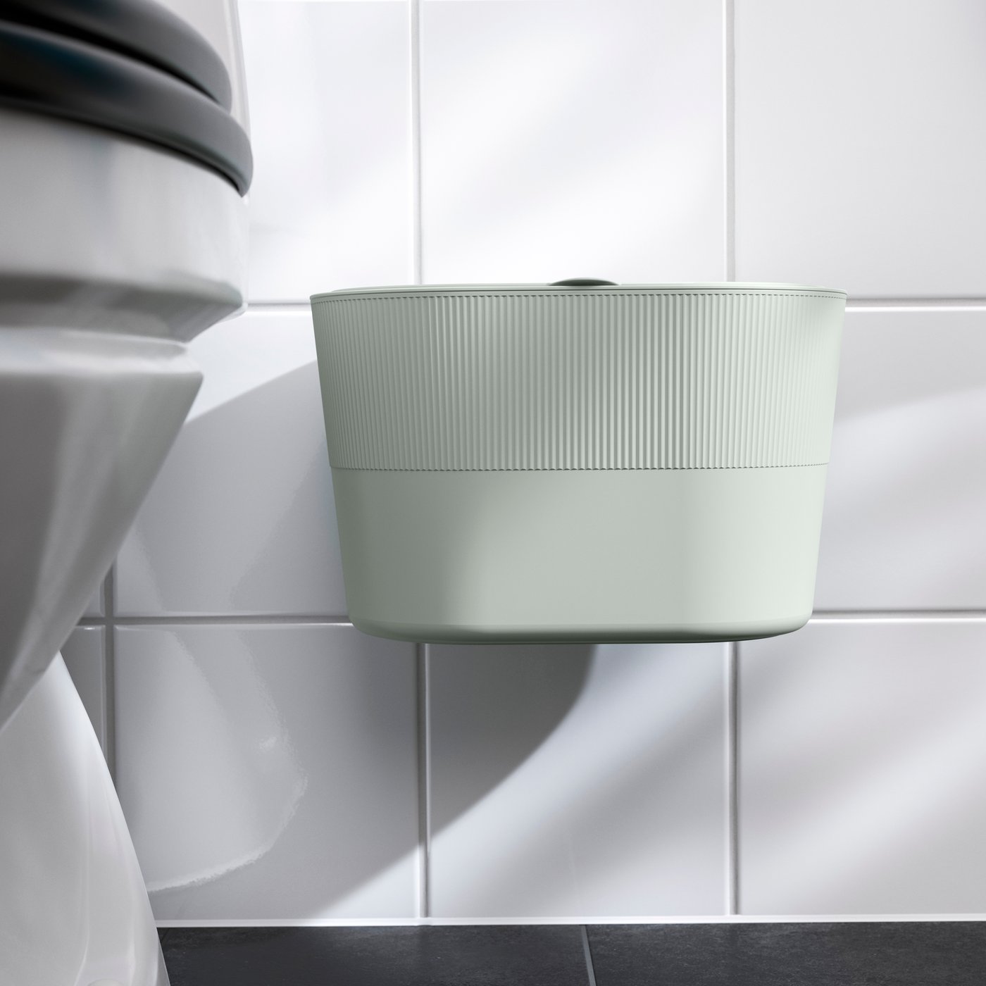 Imaginea prezintă o toaletă albă și un coș de gunoi verde deschis montat pe perete, într-o baie cu faianță albă și podea din plăci ceramice negre.