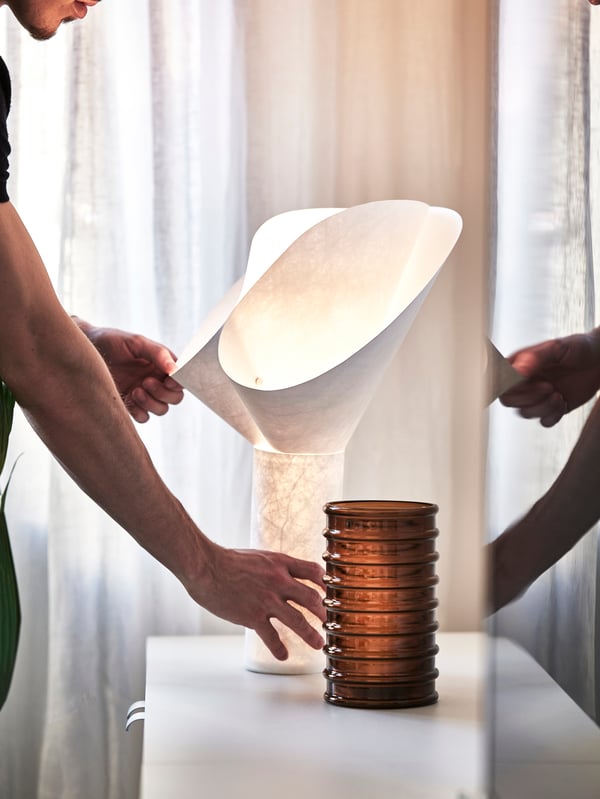 Person justerar en vit, skulptural BÄCKNATE bordslampa bredvid en staplad dekorativ behållare på ett bord framför ett fönster.