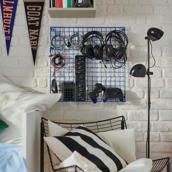 A blue GREJIG wall grid is mounted on a white brick wall, holding headphones, keyboards and video game hand controllers.